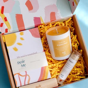 May include: A gift box with the text "For your soul" on the lid. Inside are affirmation cards, a candle that says "Smells Like Brighter Days", a tube of bath salts, and a wooden roller. The box is filled with yellow paper.