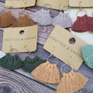 May include: Assortment of handmade tassel earrings in various colors, including brown, beige, green, and red. The earrings are displayed on brown card stock with the text "Knotted & Known" printed on each card.