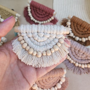 Puede incluir: Un conjunto de seis pequeñas tapices de macramé de pared en varios colores, incluyendo beige, marrón, rosa y rojo. Cada tapiz presenta una forma de semicírculo con un borde de flecos y cuentas de madera.