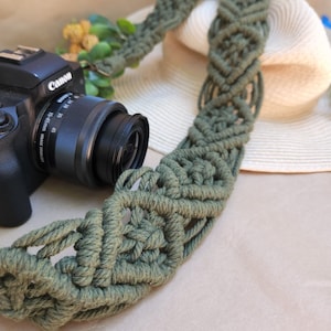 Puede incluir: Una correa de cámara de macramé verde con una cámara Canon y un sombrero de paja blanco en el fondo.