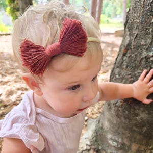 Puede incluir: Primer plano de un niño con una diadema con lazo de macramé color óxido. El lazo está centrado en una fina banda elástica de color claro. El niño lleva una camisa rosa claro y está cerca de un árbol.