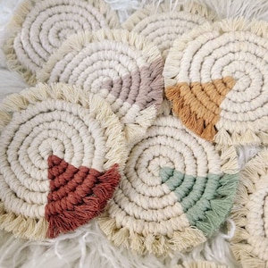May include: A collection of handmade macrame coasters. Each coaster features a spiral design in cream-colored rope, with a colorful triangle accent in shades of rust, tan, sage green, and mauve. The coasters have a fringed edge.