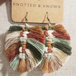 May include: A pair of knotted and woven tassel earrings in shades of green, brown, cream, and rust. The earrings are made with a variety of colors and textures, and they are perfect for adding a touch of bohemian style to any outfit.