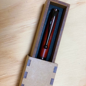 Op de afbeelding: Een houten pen met een donkerbruine en rode houtnerf, in een bruine kartonnen doos met een zwarte binnenkant.