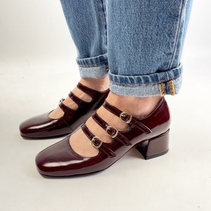 May include: Burgundy patent leather Mary Jane shoes with three straps and gold buckles. The shoes have a square toe and a block heel. The shoes are worn with blue jeans.