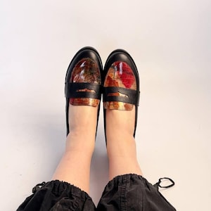Puede incluir: Un par de mocasines de cuero negro con una pala estampada y colorida. Los mocasines tienen una correa en la parte superior y est&aacute;n sobre un fondo blanco. El estampado de la pala incluye tonos rojos, marrones y dorados.