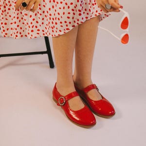 May include: A pair of bright red Mary Jane shoes with a smooth finish, featuring a rounded toe, a scalloped edge along the opening, and a single strap across the instep secured by a decorative silver-toned buckle with small clear inset stones.