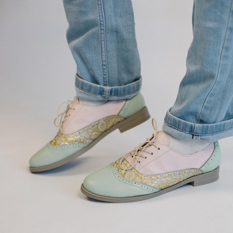 K&ouml;nnte beinhalten: Ein Paar Schn&uuml;rschuhe im Oxford-Stil mit einem mehrfarbigen Design aus Pastellrosa, Mintgr&uuml;n und einem goldenen Blumenmuster-Einsatz. Die Schuhe haben eine abgerundete Spitze, einen niedrigen Absatz und d&uuml;nne beige Schn&uuml;rsenkel, getragen zu hellblauen Jeans.