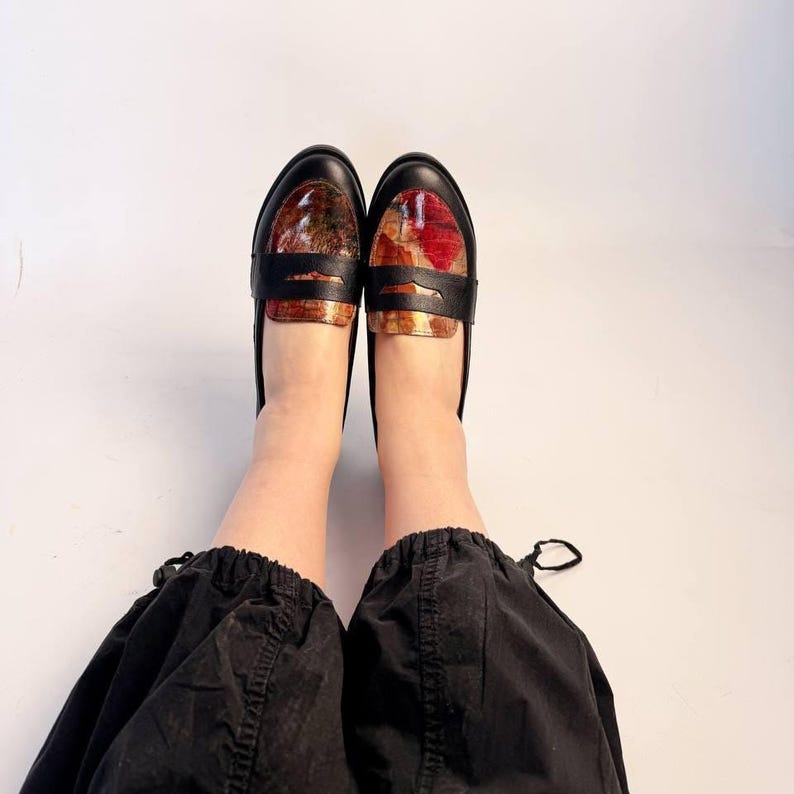 Puede incluir: Un par de mocasines de cuero negro con una pala estampada y colorida. Los mocasines tienen una correa en la parte superior y est&aacute;n sobre un fondo blanco. El estampado de la pala incluye tonos rojos, marrones y dorados.