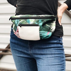 Könnte beinhalten: Eine Bauchtasche mit tropischem Blättermuster in Grün-, Blau- und Rosatönen und einer weißen, lederartigen Vordertasche. Die Tasche hat einen schwarzen Reißverschluss und einen schwarzen, verstellbaren Riemen.