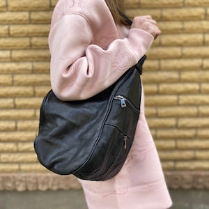Peut inclure: Un sac bandoulière en cuir noir avec un design incurvé et une fermeture éclair. Le sac est porté sur l'épaule contre un sweat-shirt rose clair. L'arrière-plan est un mur de briques brun clair texturé.
