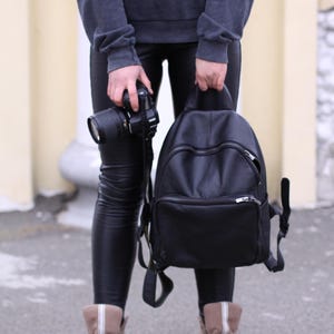 May include: A black leather backpack featuring a rounded top, a large main compartment with a zipper closure, and a smaller front pocket with a horizontal zipper. The backpack has adjustable shoulder straps and silver-toned metal hardware.