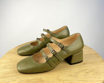 May include: Olive green Mary Jane style shoes with three straps and silver buckles. The shoes have a square toe and a block heel. The shoes are displayed on a light brown wooden surface.