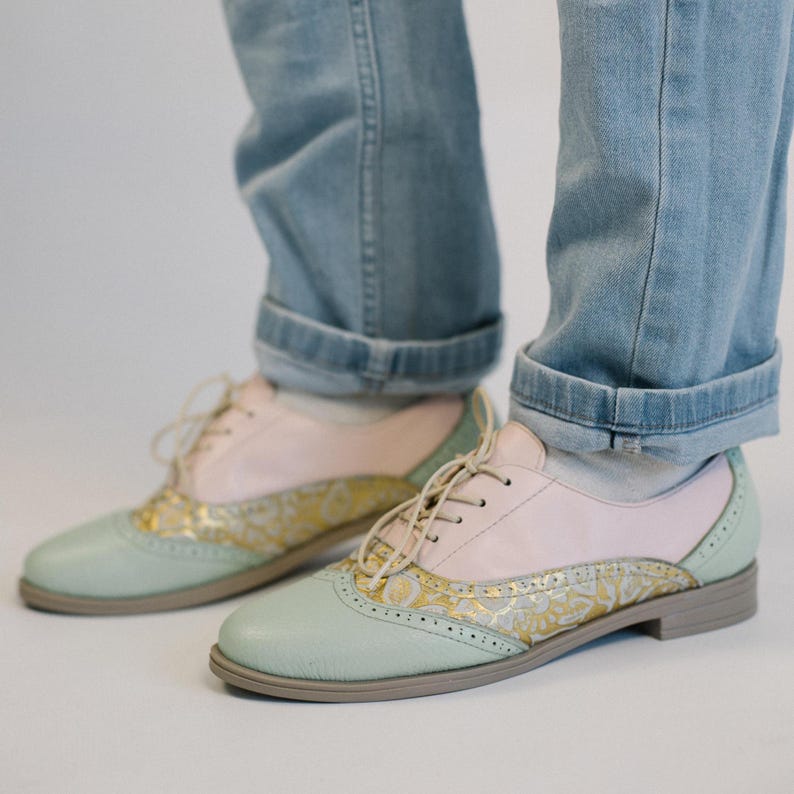 K&ouml;nnte beinhalten: Ein Paar Schn&uuml;rschuhe im Oxford-Stil mit hellrosa Obermaterial, mintgr&uuml;ner Zehenkappe und Fersenkappe sowie einem goldfarbenen Seitenteil mit Blumenmuster. Die Schuhe haben einen flachen Absatz und werden zu hellblauen Jeans mit Umschlag und wei&szlig;en Socken getragen.