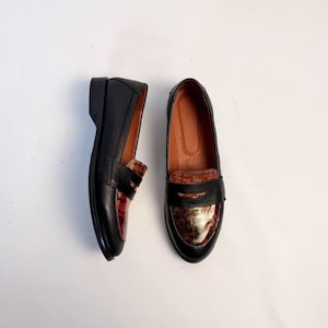 Puede incluir: Un par de mocasines de cuero negro con una correa decorativa en la parte superior. La puntera y el tal&oacute;n presentan un patr&oacute;n texturizado marr&oacute;n rojizo. Los zapatos tienen un tac&oacute;n bajo y un dise&ntilde;o cl&aacute;sico.