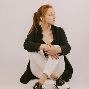 May include: A pair of black and white saddle shoes with black laces, featuring a rounded toe, a low heel, and a classic two-tone leather design. The shoes are worn with white trousers and a black long-sleeved button-down shirt.