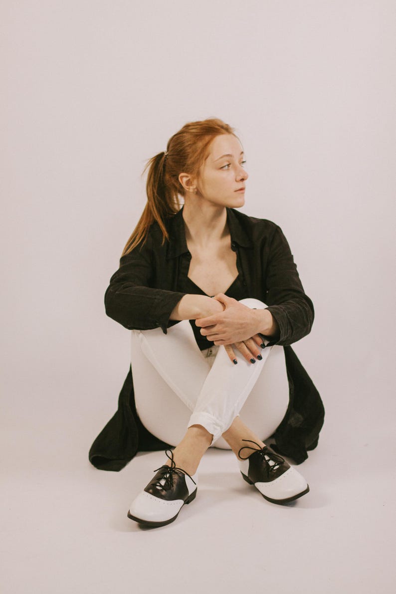 May include: A pair of black and white saddle shoes with black laces, featuring a rounded toe, a low heel, and a classic two-tone leather design. The shoes are worn with white trousers and a black long-sleeved button-down shirt.