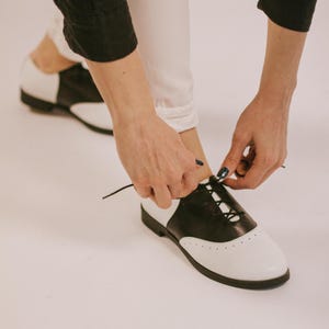 Handmade black and white saddle shoes crafted from genuine leather with a classic two-tone Oxford design. Vintage-inspired lace-up flats featuring a comfortable low-profile sole, soft leather lining, and timeless spectator style. Perfect for everyday wear, swing dancing, rockabilly fashion, retro outfits, vintage wardrobes, teachers, office looks, and casual styling.