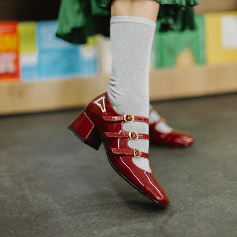 May include: Glossy red Mary Jane style shoes with three straps and gold buckles. The shoes have a block heel and a pointed toe. The shoes are worn with gray socks.