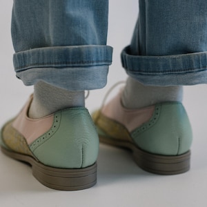 K&ouml;nnte beinhalten: Ein Paar Oxford-Schuhe mit mehrfarbigem Design, bestehend aus pastellgr&uuml;nen Fersenkappen, hellrosa Seitenteilen und gelb gemusterten Zehenkappen. Die Schuhe haben wei&szlig;e Schn&uuml;rsenkel, hellgraue Socken und braune Sohlen mit flachem Absatz.
