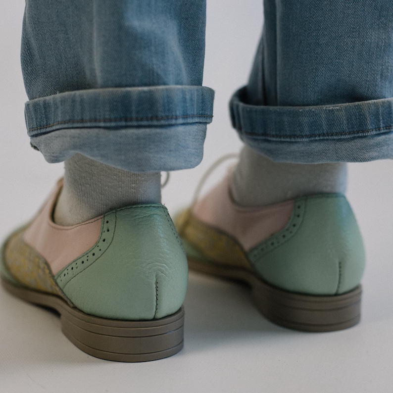 K&ouml;nnte beinhalten: Ein Paar Oxford-Schuhe mit mehrfarbigem Design, bestehend aus pastellgr&uuml;nen Fersenkappen, hellrosa Seitenteilen und gelb gemusterten Zehenkappen. Die Schuhe haben wei&szlig;e Schn&uuml;rsenkel, hellgraue Socken und braune Sohlen mit flachem Absatz.