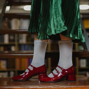 May include: Red patent leather Mary Jane shoes with three straps and a block heel. The shoes are worn with silver glitter socks and are on a wooden staircase. The background is a library with bookshelves.