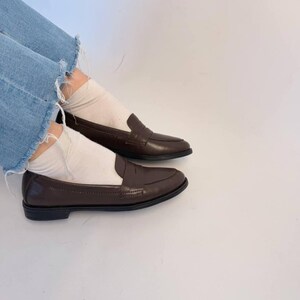 The deep brown leather color creates a sophisticated warm neutral tone suitable for classic everyday wardrobes, quiet luxury aesthetics, vintage-inspired styling, and minimalist fashion. Styled casually with cropped denim and white socks, the outfit creates a modern effortless street style look inspired by Scandinavian fashion and contemporary capsule wardrobe trends.