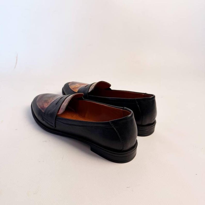 Puede incluir: Un par de mocasines de cuero negro con una pala decorativa con estampado marr&oacute;n y beige. Los zapatos tienen un dise&ntilde;o cl&aacute;sico sin cordones con un tac&oacute;n bajo y un acabado liso y pulido. Los zapatos se muestran sobre un fondo blanco.