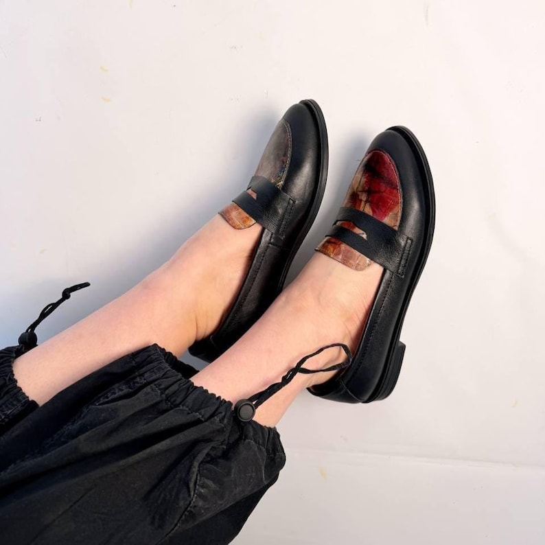 Puede incluir: Mocasines de cuero negro con correas decorativas. La puntera y la correa presentan un patr&oacute;n &uacute;nico de tonos rojos, marrones y beige. Los zapatos se muestran sobre un fondo blanco.