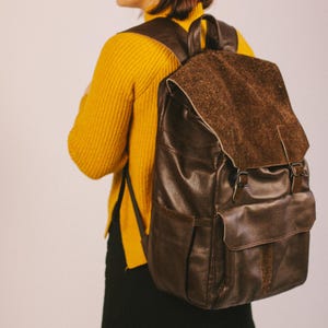 Könnte beinhalten: Ein dunkelbrauner Lederrucksack mit einer strukturierten Klappe, zwei Schnallenriemen, einer Vordertasche und Seitenfächern. Der Rucksack verfügt über verstellbare Schultergurte und einen Tragegriff sowie eine genarbte Lederoberfläche.