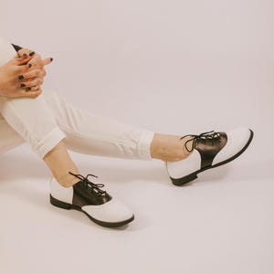 May include: A pair of black and white saddle shoes featuring a smooth leather-like finish, black laces, and a low heel. The shoes have a classic two-tone design with a white toe and heel cap, a black mid-section, and decorative perforations along the seams.