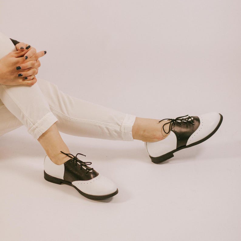 May include: A pair of black and white saddle shoes featuring a smooth leather-like finish, black laces, and a low heel. The shoes have a classic two-tone design with a white toe and heel cap, a black mid-section, and decorative perforations along the seams.