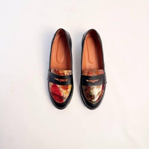 Puede incluir: Un par de mocasines de cuero negro con una pala estampada y colorida. Los mocasines tienen un dise&ntilde;o cl&aacute;sico con punta redonda y tac&oacute;n bajo. La pala presenta una mezcla de tonos rojos, naranjas y amarillos.