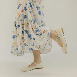 Könnte beinhalten: Cremefarbene Lederballerinas mit T-Riemen und Schnallenverschluss. Die Schuhe werden mit einem weißen Kleid mit Blumenmuster getragen. Die Ballerinas haben ein schlichtes, elegantes Design, das für verschiedene Anlässe geeignet ist.