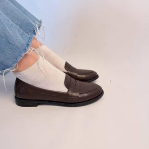 The smooth leather reflects soft studio lighting that enhances the premium quality and elegant texture of the material. The classic penny strap detail across the vamp adds timeless character while maintaining a clean minimalist aesthetic. The shoes are photographed from a side angle that clearly displays the profile, toe shape, stitching, and elegant simplicity of the design. The white background keeps attention fully on the footwear and styling details.