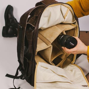 Op de afbeelding: Een donkerbruine leren camerarugzak met een getextureerde buitenkant en zwarte riemen. De binnenkant is voorzien van een beige stoffen voering met verstelbare gewatteerde verdelers en een ritsvak aan de voorflap van de rugzak.