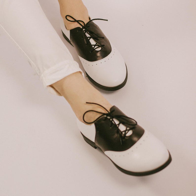 May include: A pair of black and white saddle shoes featuring a smooth leather construction, a rounded toe, and black laces. The shoes have a distinct two-tone color block design with perforated detailing along the seam of the saddle overlay.