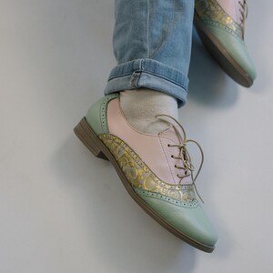 K&ouml;nnte beinhalten: Ein Paar Schn&uuml;rschuhe im Oxford-Stil mit einem Color-Blocking-Design in Pastellrosa und Minzgr&uuml;n. Die Schuhe verf&uuml;gen &uuml;ber goldene Metallic-Eins&auml;tze mit einem komplizierten Blumenmuster, Brogue-Details an den N&auml;hten und einen niedrigen braunen Absatz.