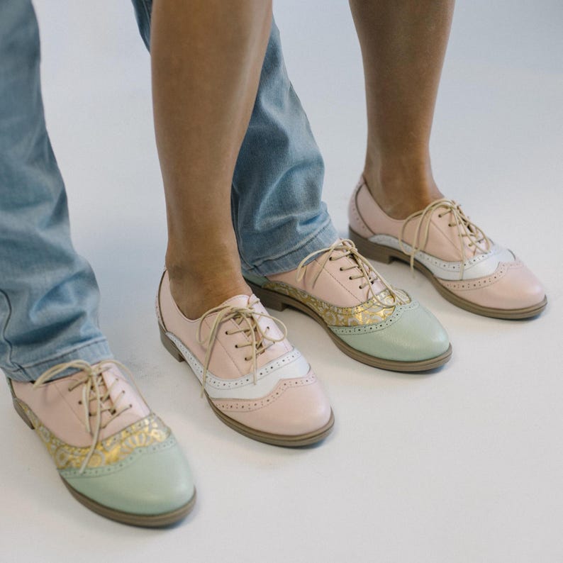 K&ouml;nnte beinhalten: Ein Paar Schn&uuml;rschuhe im Oxford-Stil mit einem mehrfarbigen Design aus pastellrosa, mintgr&uuml;nen und wei&szlig;en Eins&auml;tzen. Die Schuhe verf&uuml;gen &uuml;ber einen goldfarbenen, gemusterten Bereich an der Schuhspitze, beige Schn&uuml;rsenkel und einen flachen braunen Absatz.