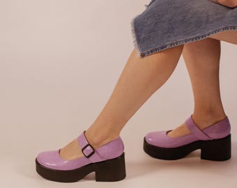 May include: A pair of lavender Mary Jane shoes with a glossy finish, featuring a rounded toe, a single strap across the instep with a black buckle, and a thick, chunky black platform sole that provides significant elevation and a solid base.