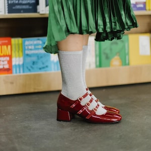 May include: Burgundy patent leather Mary Jane shoes with three straps and small block heels. The shoes are worn with silver glitter socks and a green pleated skirt. The background includes a bookshelf with colorful books.