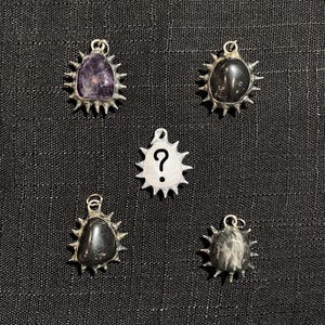 May include: Five pendants arranged in a circular pattern. Four pendants feature polished stones in purple, black, and grey tones set in silver-toned metal with radiating spikes. The center pendant is white with a black question mark symbol.