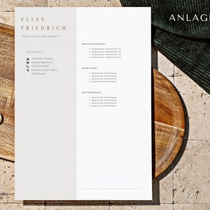 May include: A white resume with the name Elias Friedrich and the title Personalreferent. The resume includes contact information, work experience, education, and skills. The resume is on a wooden table with a green cloth in the background.