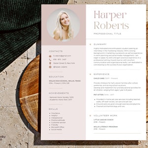 May include: A resume template with a photo of a smiling person, featuring a soft color palette. The resume includes sections for contact information, education, achievements, skills, and experience. The resume is on a wooden surface.
