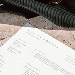 May include: A close-up of a resume with the name Calvin Norton at the top. The resume includes sections for education, work experience, and skills. The resume is printed on white paper and is lying on a textured surface.