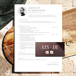 May include: A white resume with black text and a photo of a man, and a dark brown business card with the text "ATS.DE" are on a wooden tray. A gold ring is also on the tray. The resume is in German.