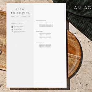May include: A minimalist resume template with the name "LISA FRIEDRICH" at the top. The document includes sections for personal details, work experience, education, and further training. The background is a light beige with a wooden tray and a gold ring.
