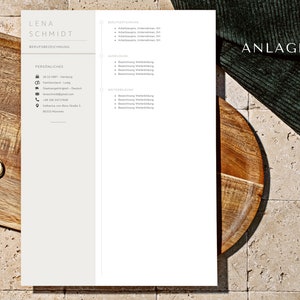May include: A resume template with the name "Lena Schmidt" at the top. The resume is on a white background with a wooden tray and a gold ring in the corner. The text "Anlagen" is visible in the top right corner.