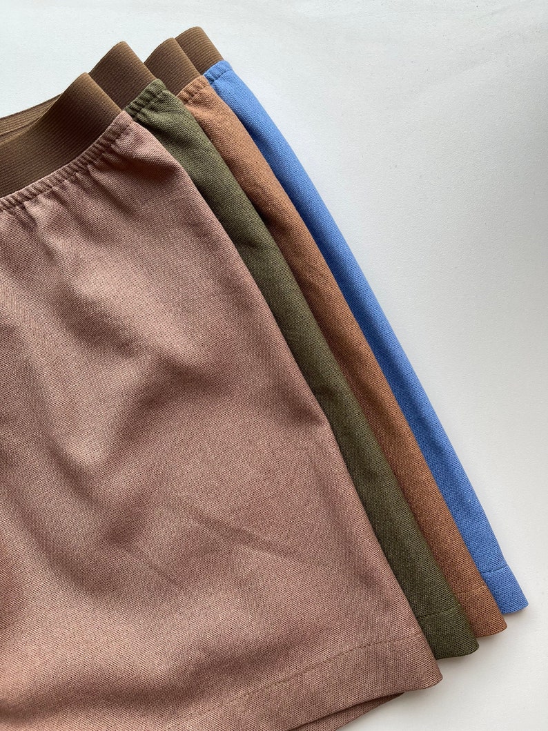 May include: A stack of four pairs of underwear in different colors: brown, olive green, brown, and light blue. The underwear is made of a soft, textured fabric.