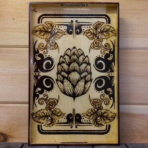 May include: A wooden serving tray with a black and white design featuring a stylized hop cone and floral motifs. The tray has a light wood background and a dark brown border.